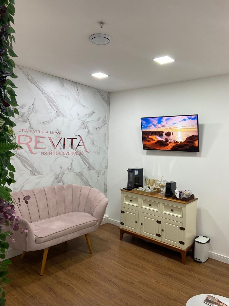 Espaço de espera com sofá rosa, parede com o nome da clínica, televisão e aparador com cantinho do café.