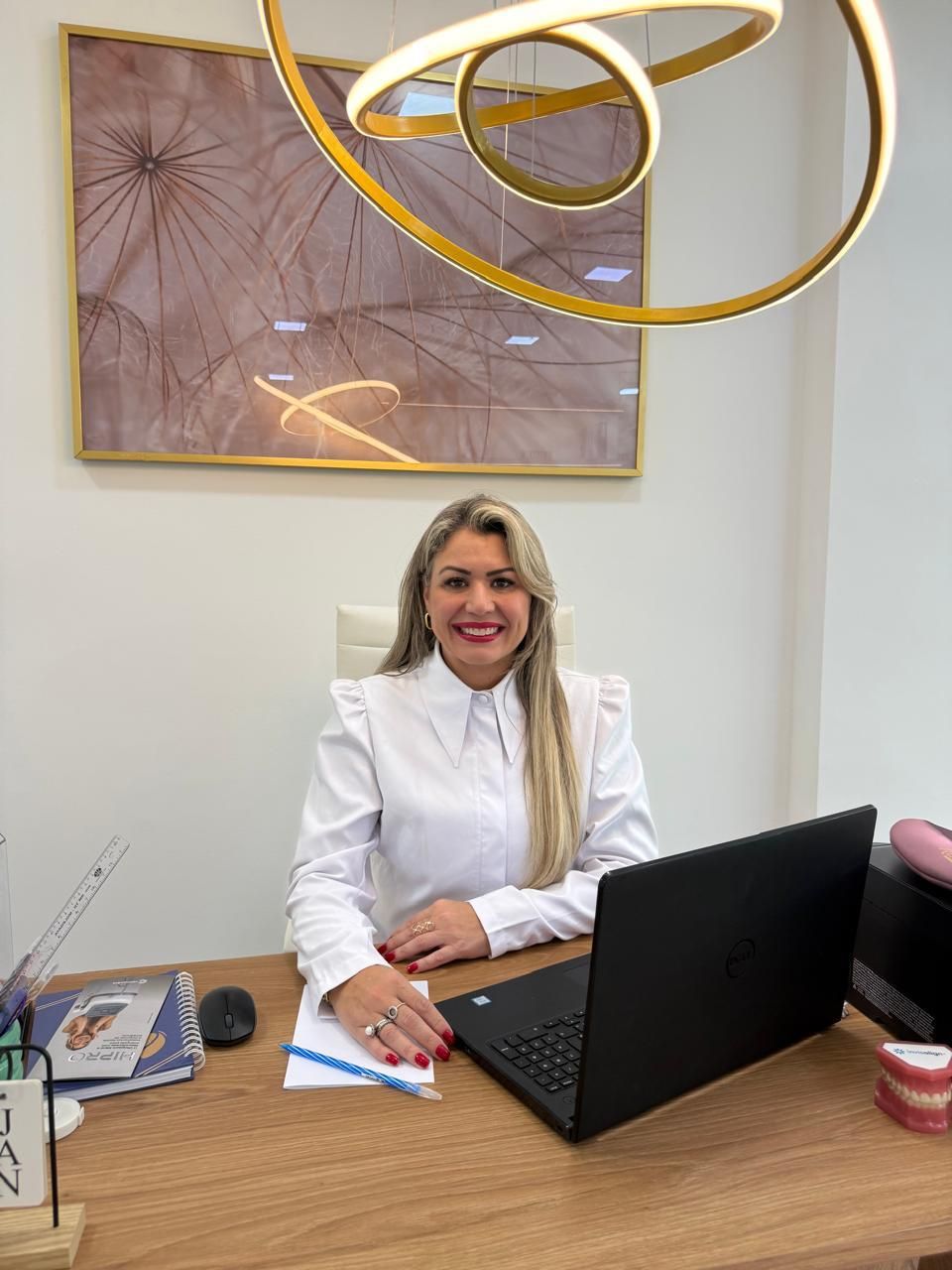 Dra. Patrícia Mussi de camisa branca, sorrindo, sentada à mesa do consultório com um notebook aberto.